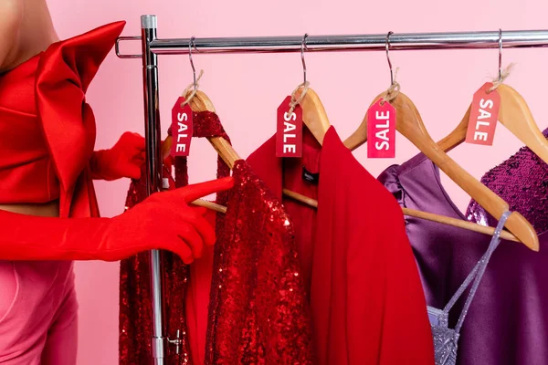Vue recadrée de la femme en gants rouges pointant vers des vêtements élégants avec des étiquettes de vente isolées sur rose — Photo de stock