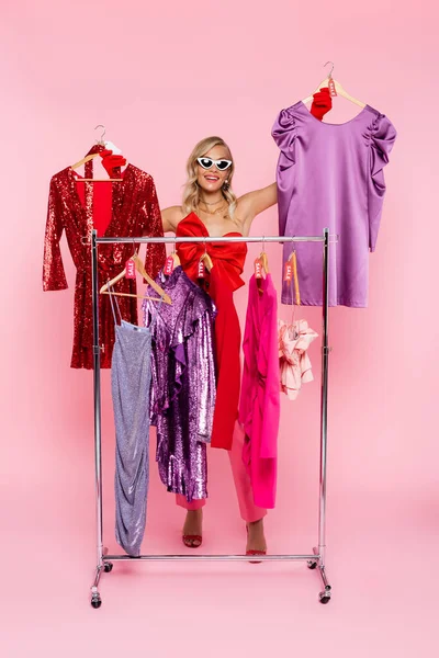 Femme joyeuse dans les lunettes de soleil tenant cintres avec des robes élégantes sur noir vendredi vente sur rose — Photo de stock