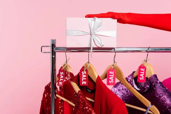 Vista ritagliata della mano femminile in guanto rosso vicino scatola regalo e rack con vestiti alla moda con tag di vendita isolati su rosa — Foto stock