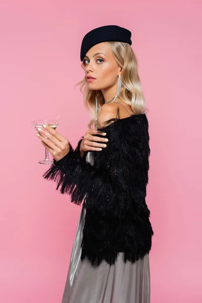 Stylish woman in black beret and faux-fur jacket looking at camera while holding cocktail isolated on pink — Stock Photo