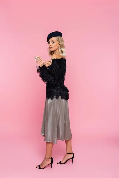 Elegant woman in black faux-fur jacket holding cocktail while looking at camera on pink — Stock Photo