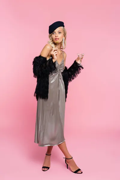 Vue pleine longueur de femme élégante en robe brillante et veste noire en fausse fourrure posant avec cocktail sur rose — Photo de stock