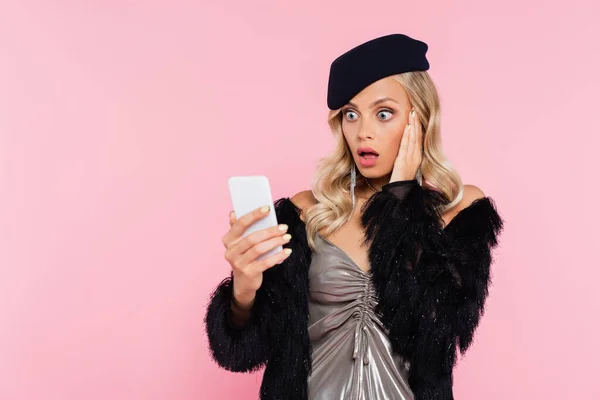 Femme étonnée dans des vêtements élégants touchant le visage tout en regardant le téléphone mobile isolé sur rose — Photo de stock