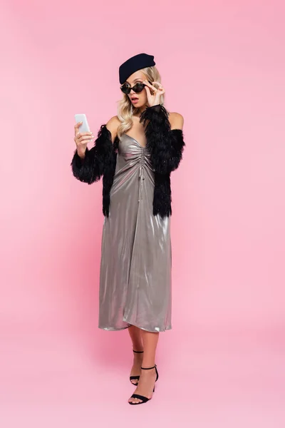 Full length view of surprised young woman in grey glossy dress and sunglasses looking at smartphone on pink — Stock Photo
