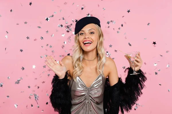Femme excitée dans des vêtements élégants tenant cocktail et agitant la main près de confettis brillants sur rose — Photo de stock