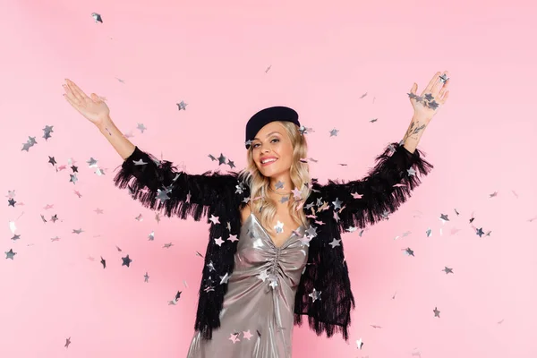 Happy woman in elegant clothes looking at camera while throwing sparkling confetti on pink — Stock Photo