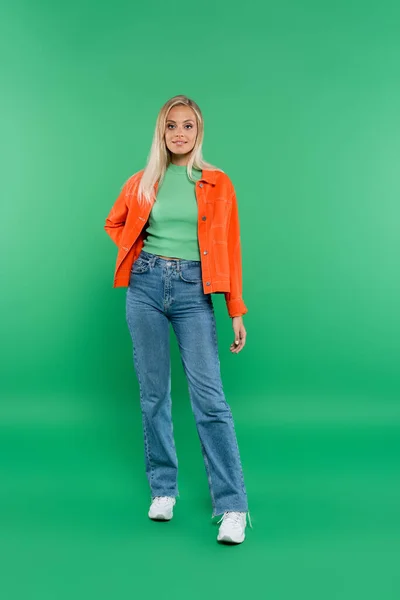 Full length view of blonde woman in jeans and orange jacket standing with hand on hip on green — Stock Photo