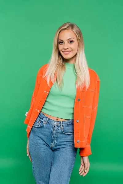 Glückliche Frau in orangefarbener Jacke und Jeans blickt in die Kamera auf grün — Stockfoto