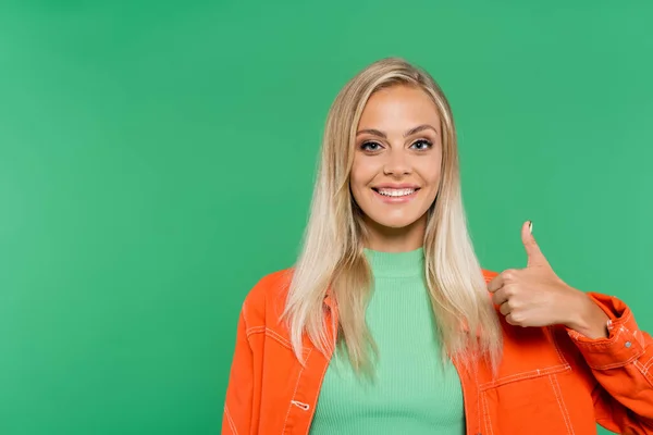 Mujer alegre en chaqueta naranja mostrando el pulgar hacia arriba mientras mira la cámara aislada en verde - foto de stock