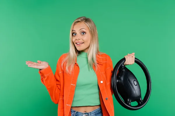 Excited blonde woman pointing with hand and holding steering wheel while looking at camera isolated on green — Stock Photo