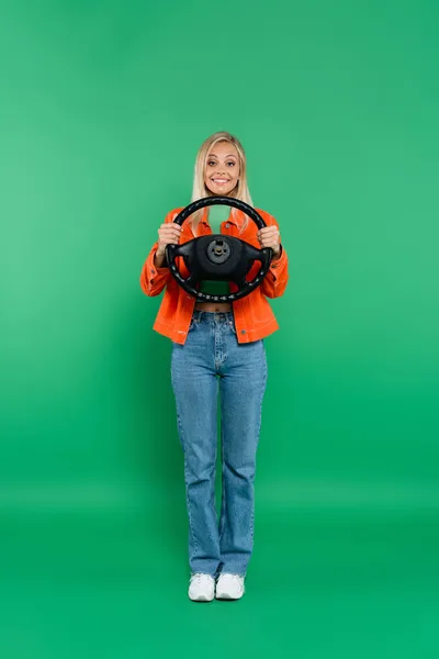 Visão de comprimento total da mulher loira satisfeita em jeans posando com volante no verde — Fotografia de Stock