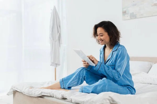 Jovem alegre em pijama azul usando tablet digital no quarto — Fotografia de Stock