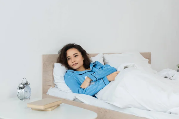 Брюнетка женщина лежит в постели возле прикроватного столика с книгами и будильником — стоковое фото