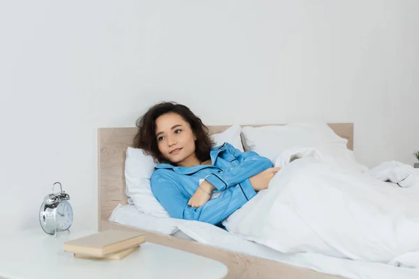 Morena tumbada en la cama cerca de la mesita de noche con libros y despertador - foto de stock