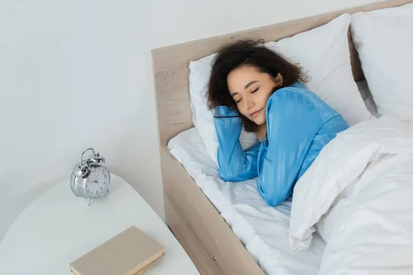 Vista ad alto angolo della donna bruna che dorme nel letto vicino al comodino con libri e sveglia — Foto stock