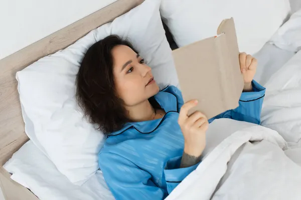 Высокий угол зрения брюнетки, читающей книгу в постели — стоковое фото