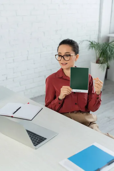 Young teacher in eyeglasses holding card with green lettering and looking at laptop — Stock Photo