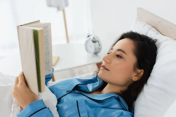 Mujer joven en pijama azul leyendo libro en la cama - foto de stock