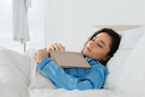 Jeune femme en pyjama bleu dormant avec livre au lit — Photo de stock