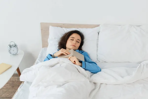 Brunetta donna in pigiama blu dormire con libro a letto — Foto stock