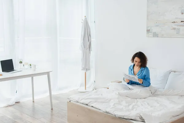 Morena mujer en pijama azul leyendo la vida de viaje periódico en la cama cerca del ordenador portátil en el escritorio - foto de stock