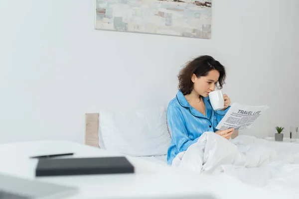 Brunette woman in blue pajamas reading travel life newspaper and drinking tea in bed — Stock Photo