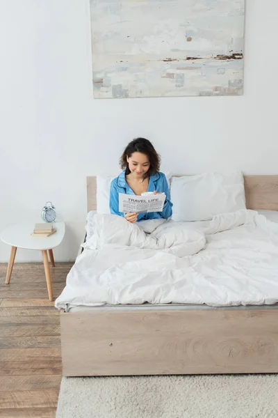 Fröhliche junge Frau im Schlafanzug liest Reisezeitung und hält Tasse im Bett — Stockfoto