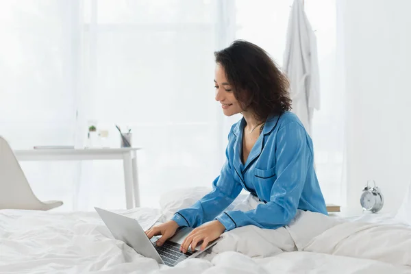 Jovem freelancer alegre em pijama usando laptop na cama — Fotografia de Stock