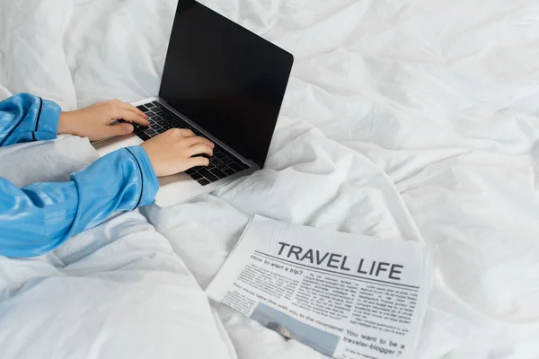 Vista cortada de jovem em pijama usando laptop perto de jornal na cama — Fotografia de Stock