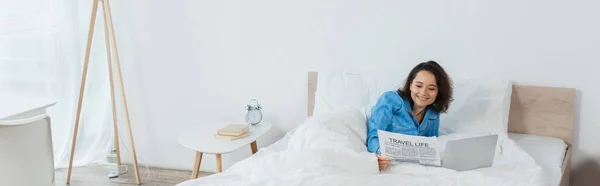 Joyeuse jeune femme en pyjama lire journal près d'un ordinateur portable sur le lit, bannière — Photo de stock