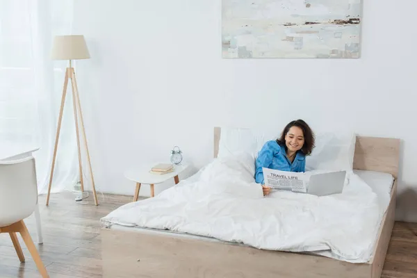 Fröhliche junge Frau im Schlafanzug liest Zeitung neben Laptop auf dem Bett — Stockfoto