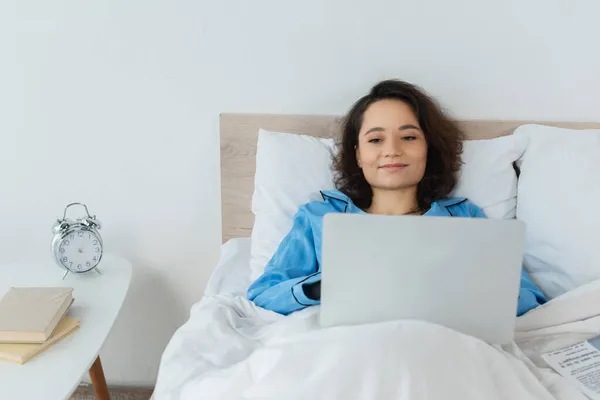 Freelancer jovem positivo em pijama azul usando laptop na cama — Fotografia de Stock