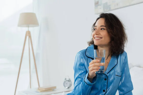 Счастливая молодая женщина в голубой пижаме держит стакан воды в спальне — стоковое фото