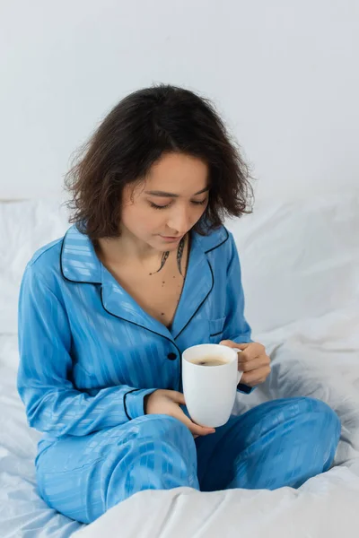 Jeune femme en pyjama bleu tenant une tasse de café dans la chambre — Photo de stock