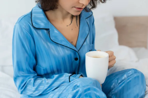 Cropped view of tattooed young woman in blue pajamas holding cup of coffee in bedroom — Stock Photo