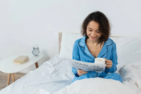 Jovem alegre em pijama lendo viagem vida jornal e segurando copo na cama — Fotografia de Stock