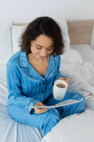 Vista ad alto angolo di giovane donna in pigiama blu con in mano una tazza di caffè mentre legge il giornale in camera da letto — Foto stock