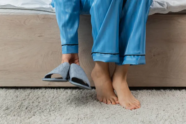 Cropped view of young woman in blue pajamas holding slippers in bedroom — Stock Photo