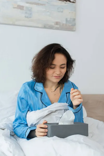 Mujer enferma y tatuada estornudando cerca de la caja de tejido en la cama - foto de stock