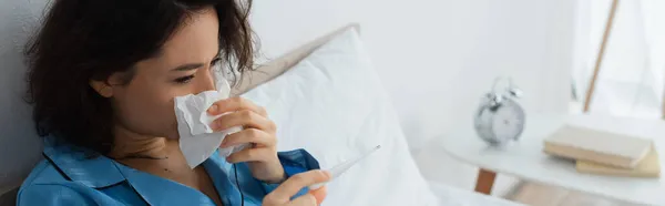 Femme malade éternuer dans les tissus et en regardant thermomètre électronique, bannière — Photo de stock