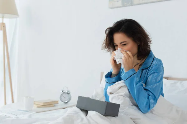 Femme malade éternuer dans la serviette près de la boîte de tissu sur le lit — Photo de stock