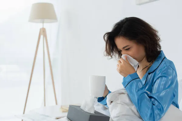 Sick woman sneezing in napkin while holding cup of tea — Stock Photo