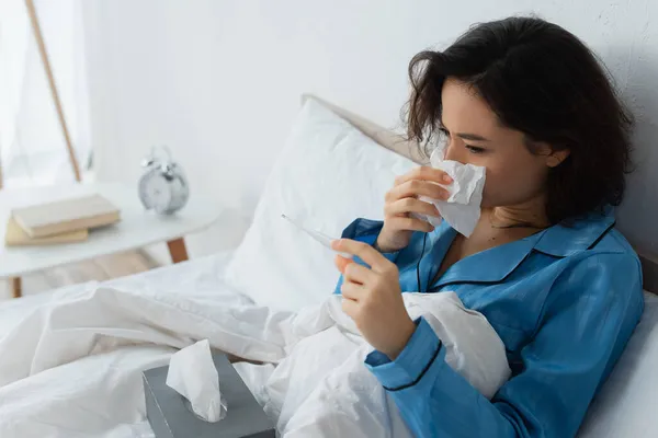 Sick woman sneezing in tissue and looking at electronic thermometer — Stock Photo