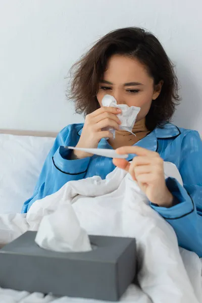 Mujer joven enferma estornudando en tejido y mirando el termómetro electrónico - foto de stock
