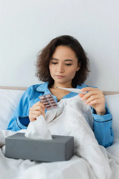 Хвора молода жінка тримає блістерний пакет з таблетками та електронним термометром — стокове фото