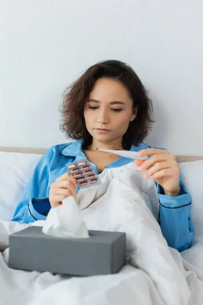 Jovem doente segurando blister pack com pílulas e termômetro eletrônico — Fotografia de Stock