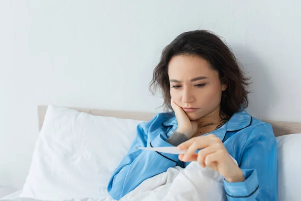 Kranke junge Frau blickt auf elektronisches Thermometer — Stockfoto