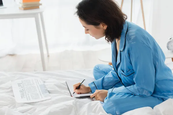 Mujer joven escribiendo en cuaderno cerca de periódico en la cama - foto de stock