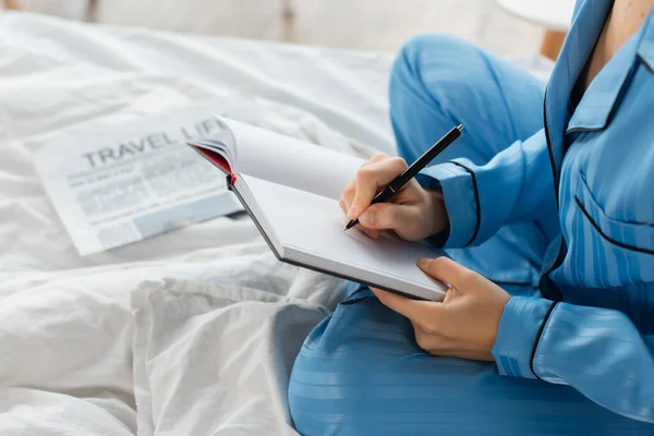 Vista recortada de la joven mujer escribiendo en cuaderno cerca de periódico en la cama - foto de stock
