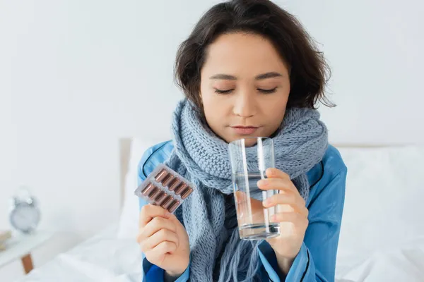 Sick young woman in knitted scarf holding glass of water and blister pack with pills — Stock Photo